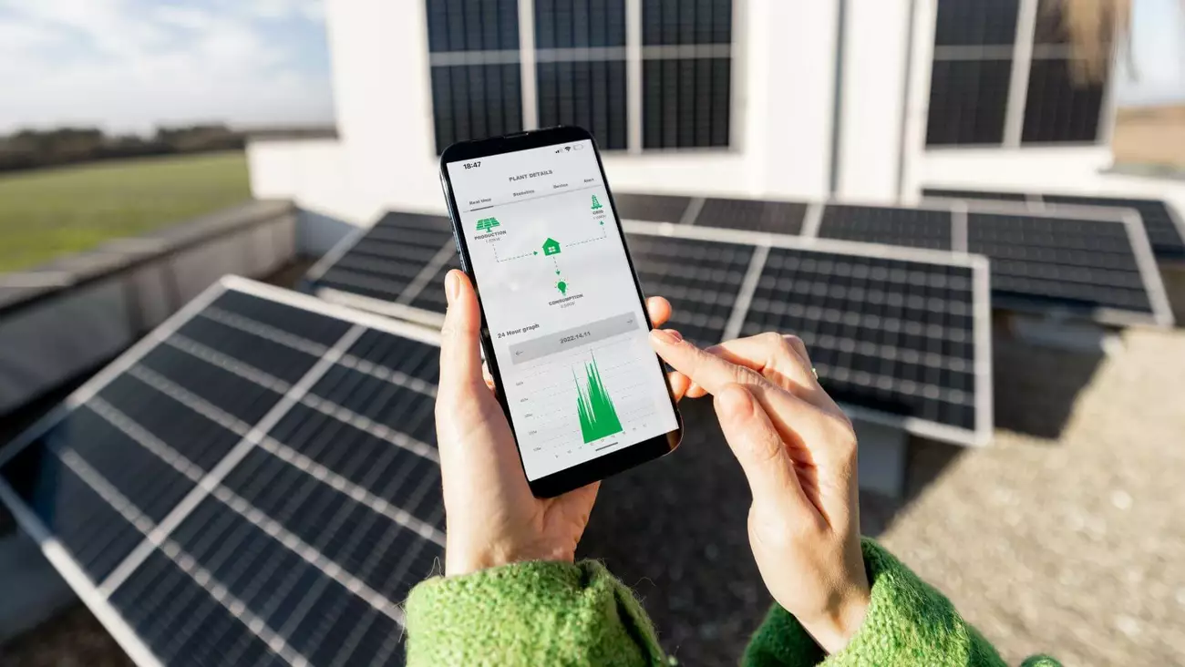 Person hält Smartphone mit Energieverbrauchs-App vor Solarpanels. Auf dem Display sind Echtzeit-Daten zum Stromverbrauch und Preisverlauf zu sehen.