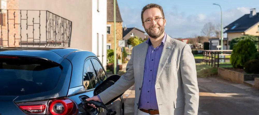 Ein lächelnder Mann mit Brille, hellem Sakko, lila Hemd und weißer Hose steht draußen auf einer Wohnstraße neben einem schwarzen Elektroauto und steckt ein Ladekabel ein. Im Hintergrund sind Wohnhäuser, ein Gartenzaun und eine Straßenlaterne zu sehen.