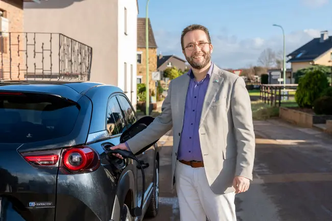 Ein lächelnder Mann mit Brille, hellem Sakko, lila Hemd und weißer Hose steht draußen auf einer Wohnstraße neben einem schwarzen Elektroauto und steckt ein Ladekabel ein. Im Hintergrund sind Wohnhäuser, ein Gartenzaun und eine Straßenlaterne zu sehen.